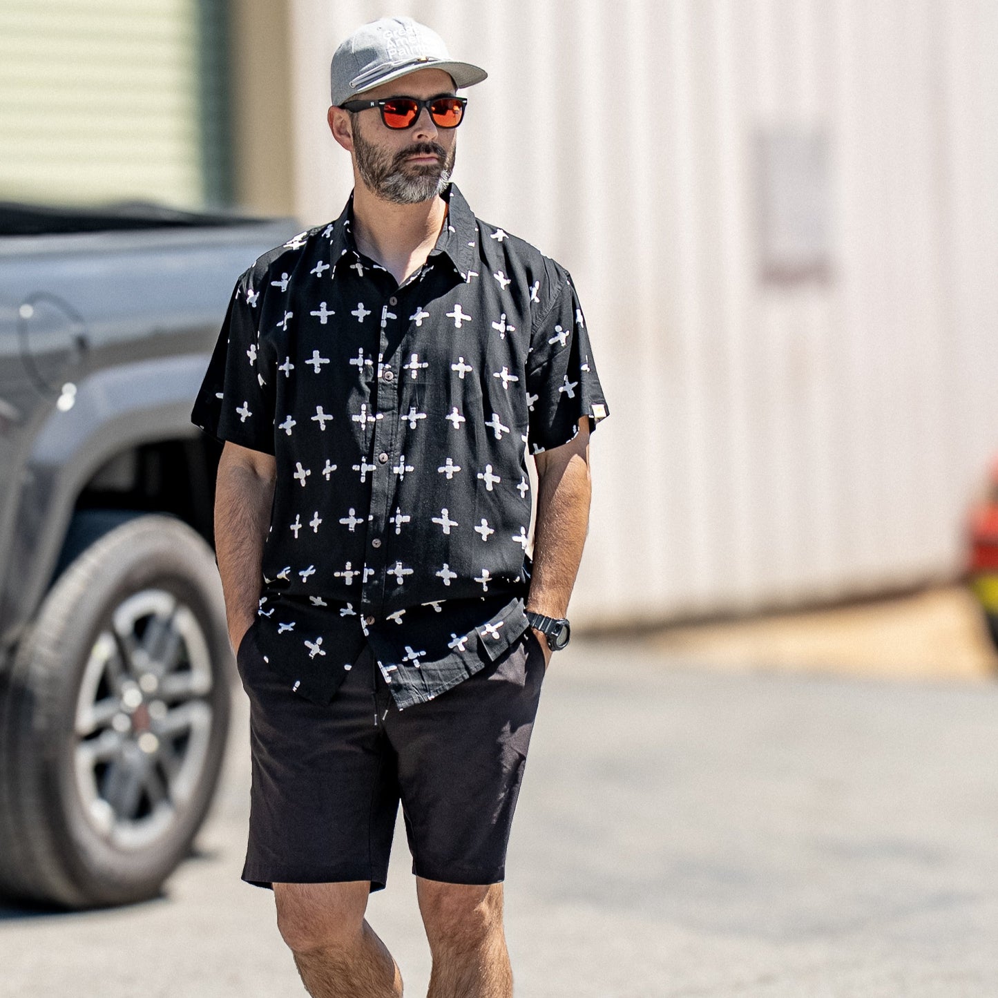 Man wearing a Hormesis attiere black crux shirt, sunglasses, and shorts walking outdoors. Short Sleeve Collared shirt style. Fashion